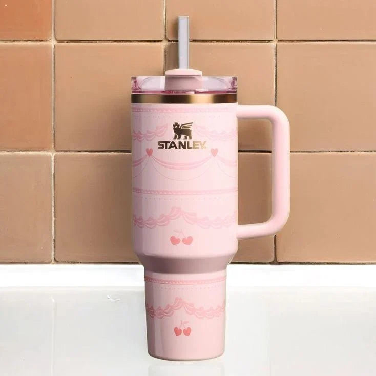 A pink Stanley travel mug with a handle, a clear lid with a white straw, and rose gold trim, set against a background of tan ceramic tiles.