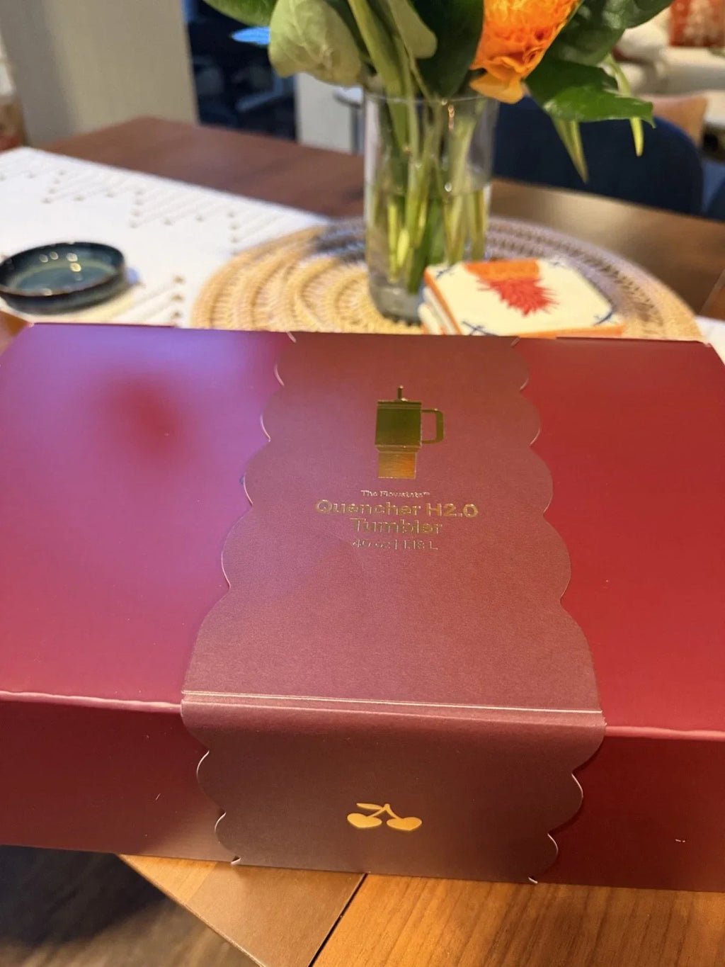 A red rectangular gift box with a scalloped flap closure and gold printed text reading 'Guatemalan H2.0 Tumbler Hot Tea' sits on a wooden table.