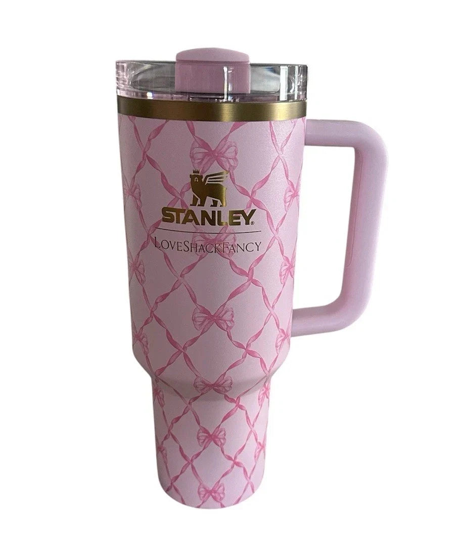 A pink Stanley tumbler with a handle, featuring a pink bow and chain-link pattern, a gold Stanley logo, and a clear lid with a pink insert, shown on a white background.