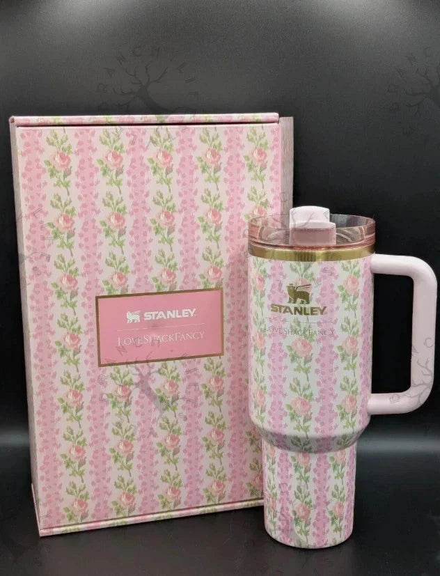 A pink floral Stanley tumbler with a white handle and lid, shown next to its matching pink patterned gift box on a dark background.