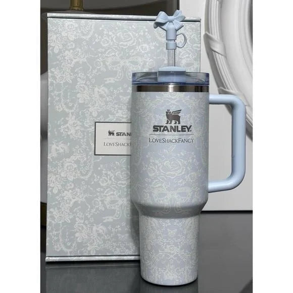 A light blue Stanley Love Shack Fancy insulated tumbler with a matching lid and handle, shown in front of its patterned gift box on a dark surface.