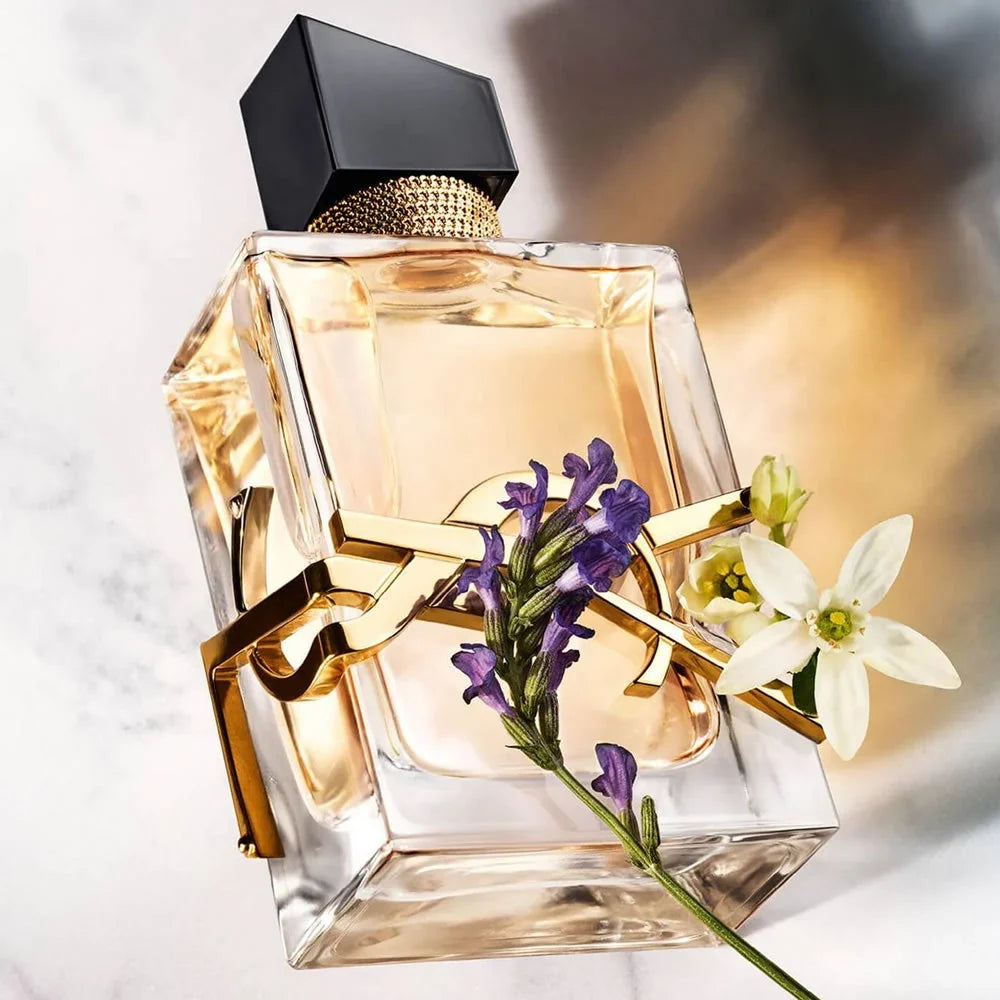 A luxury perfume bottle with a clear glass body, amber liquid, a black cap, and a gold YSL logo band, decorated with purple lavender and white flowers against a soft marble background.