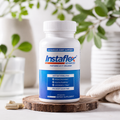 A white bottle of Instaflex Advanced dietary supplement with a blue label, displayed on a wooden slice coaster with two capsules nearby, set against a soft-focus background of white vases and green leaves.