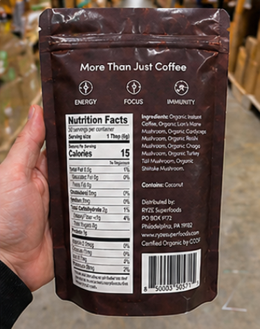 A hand holding the back of a dark brown stand-up coffee pouch, showing the nutrition facts panel, ingredients list, and barcode against an outdoor blurred background.